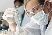 A blond medical or scientific researcher or doctor using looking at a clear solution in a laboratory with her Asian female colleague out of focus behind her.